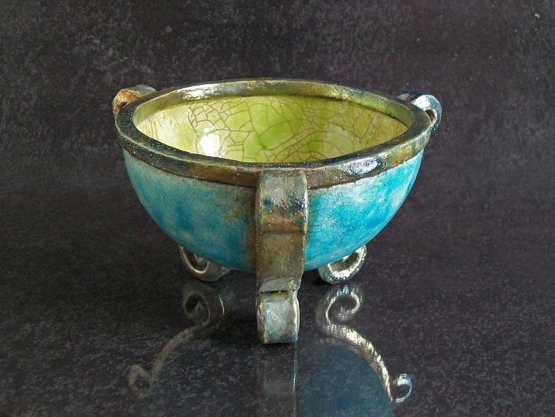 May include: A handcrafted ceramic bowl with a turquoise exterior and a lime green interior. The bowl has three small feet and two handles. The rim is a dark brown color. The bowl is approximately 6 inches in diameter.