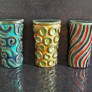 May include: Three decorative ceramic vases. The first is teal with silver swirls, the second is yellow with green circles, and the third has red and black diagonal stripes. Each vase has a metallic rim.