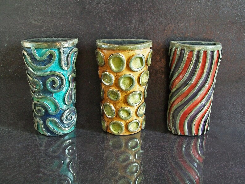May include: Three decorative ceramic vases. The first is teal with silver swirls, the second is yellow with green circles, and the third has red and black diagonal stripes. Each vase has a metallic rim.