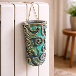 May include: A decorative, cylindrical vase with a teal and blue swirling design. The vase is suspended by a thin, tan rope. The vase is hanging on a white radiator. The vase has a metallic rim.