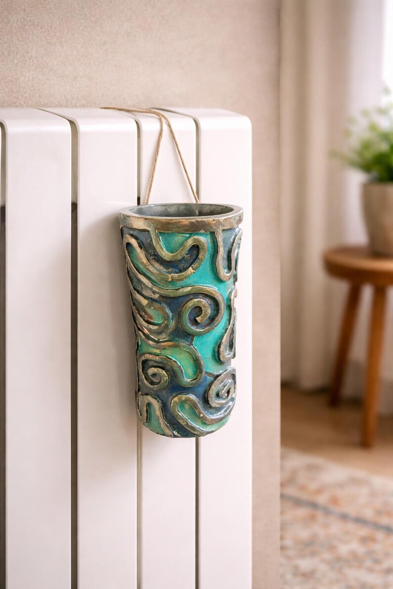May include: A decorative, cylindrical vase with a teal and blue swirling design. The vase is suspended by a thin, tan rope. The vase is hanging on a white radiator. The vase has a metallic rim.