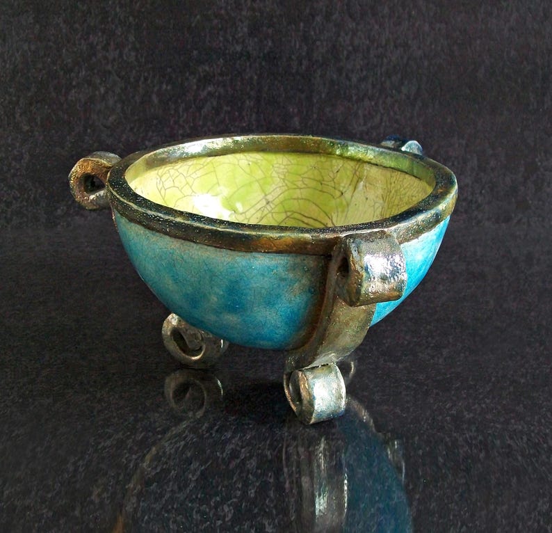 May include: A handcrafted ceramic bowl with a turquoise exterior and a lime green interior. The bowl has three small feet and two handles. The rim is accented with a gold-colored trim. The bowl is likely used for decorative purposes.