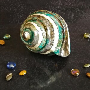 Puede incluir: Una gran concha decorativa con un patr&oacute;n en espiral de rayas turquesas, blancas, grises y marrones. La concha est&aacute; rodeada de peque&ntilde;os guijarros de vidrio de colores sobre una superficie oscura y texturizada. La concha tiene una forma natural y org&aacute;nica.