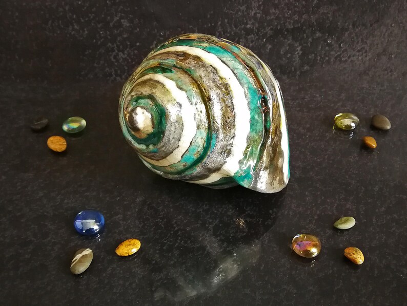 Puede incluir: Una gran concha decorativa con un patr&oacute;n en espiral de rayas turquesas, blancas, grises y marrones. La concha est&aacute; rodeada de peque&ntilde;os guijarros de vidrio de colores sobre una superficie oscura y texturizada. La concha tiene una forma natural y org&aacute;nica.