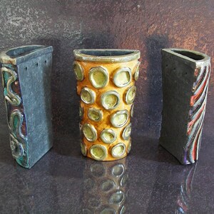 May include: Three ceramic vases with unique designs. The central vase is cylindrical with a yellow and green pattern. The flanking vases are rectangular, one with a swirling design and the other with vertical stripes. All have dark gray exteriors.