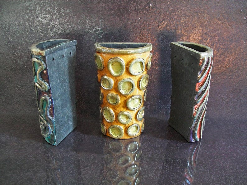 May include: Three ceramic vases with unique designs. The central vase is cylindrical with a yellow and green pattern. The flanking vases are rectangular, one with a swirling design and the other with vertical stripes. All have dark gray exteriors.