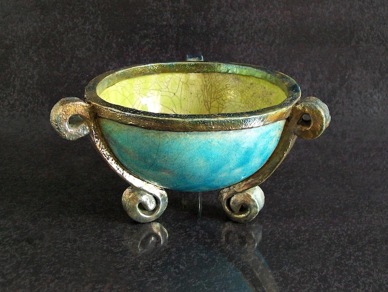 May include: A handcrafted, decorative bowl with a turquoise exterior and a yellow interior. The bowl features ornate handles and rests on three scrolled feet. The rim is accented with a gold trim, adding a touch of elegance to the ceramic piece.