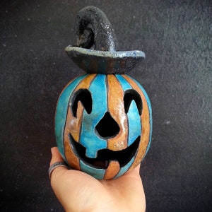 May include: A ceramic jack-o'-lantern featuring alternating vertical stripes of bright turquoise and earthy brown glaze. The pumpkin has a carved face with triangular eyes and a wide, toothy grin, topped with a textured, dark charcoal-colored witch hat.