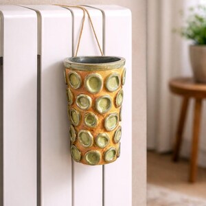 May include: A cylindrical ceramic vase with a textured surface and a pattern of raised, circular accents. The vase is primarily yellow-orange with green accents and a dark gray rim. It hangs from a beige string.