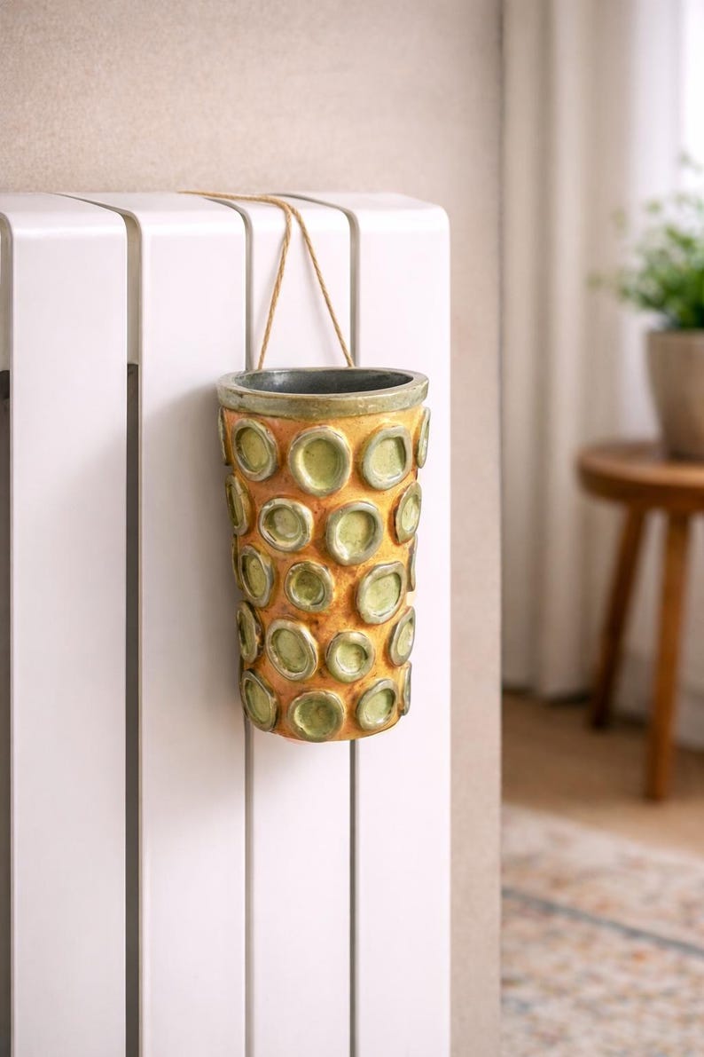 May include: A cylindrical ceramic vase with a textured surface and a pattern of raised, circular accents. The vase is primarily yellow-orange with green accents and a dark gray rim. It hangs from a beige string.