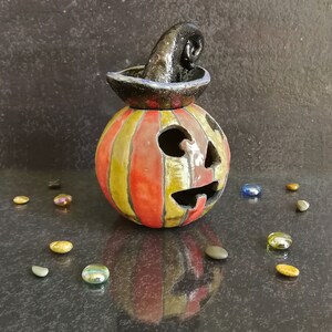 May include: A ceramic jack-o'-lantern with vertical red and yellow stripes, featuring a carved face with triangular eyes and a jagged mouth. A black, textured, curled witch hat sits on top of the pumpkin, which is surrounded by small glass stones.