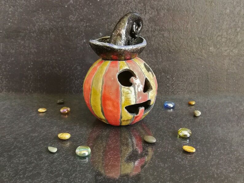 May include: A ceramic jack-o'-lantern with vertical red and yellow stripes, featuring a carved face with triangular eyes and a jagged mouth. A black, textured, curled witch hat sits on top of the pumpkin, which is surrounded by small glass stones.