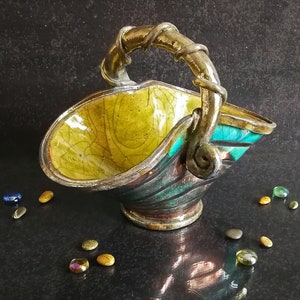 May include: A decorative basket with a gold-coloured handle and rim. The interior is yellow with a crackle pattern, and the exterior is teal and silver. Small, colourful stones surround the basket.