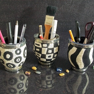 May include: Three decorative ceramic pen holders with various patterns: black circles, black and white squares, and black and white stripes. Each holder contains pens, pencils, a paintbrush, and scissors. Colorful stones are scattered around the holders.