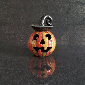 May include: A ceramic jack-o'-lantern figurine featuring vertical orange and yellow stripes with a glossy finish. The pumpkin has a carved face with triangular eyes, a nose, and a jagged mouth, topped with a black, curled, hat-like stem.