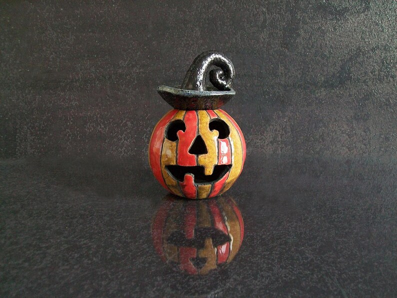 May include: A ceramic jack-o'-lantern figurine featuring vertical orange and yellow stripes with a glossy finish. The pumpkin has a carved face with triangular eyes, a nose, and a jagged mouth, topped with a black, curled, hat-like stem.