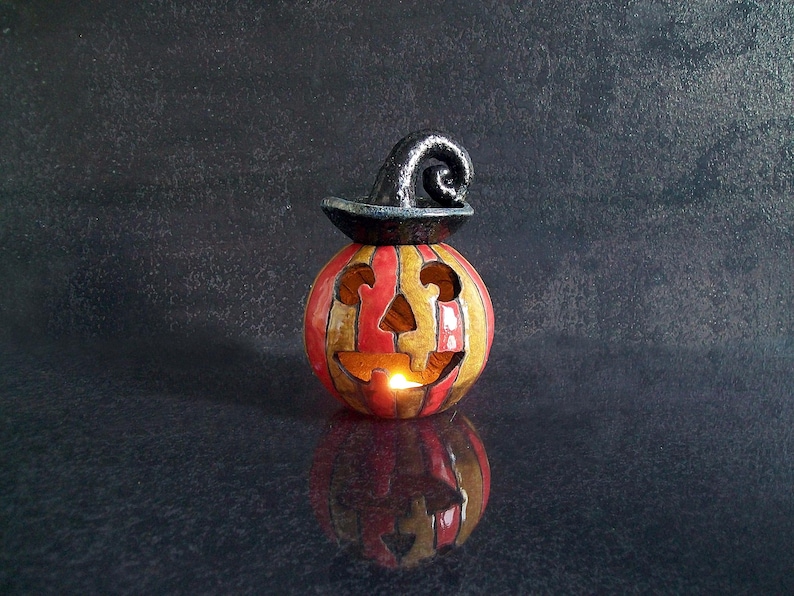 May include: A ceramic jack-o'-lantern candle holder featuring vertical red and yellow stripes, a carved face with cutouts for eyes, nose, and mouth, and a glossy black witch hat topper. A small light glows from within the hollow pumpkin body.