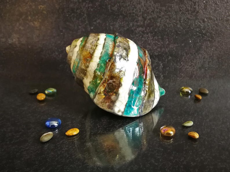 Puede incluir: Concha decorativa de cer&aacute;mica con acabado brillante. La concha presenta un patr&oacute;n a rayas en blanco, turquesa, marr&oacute;n y dorado. Peque&ntilde;as cuentas de vidrio de colores rodean la concha. Un art&iacute;culo de decoraci&oacute;n del hogar &uacute;nico.