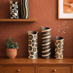 May include: A collection of decorative ceramic vases with unique patterns. The vases feature designs in black, white, and brown, including geometric shapes, stripes, and organic swirls. Some vases hold small branches with delicate white flowers.