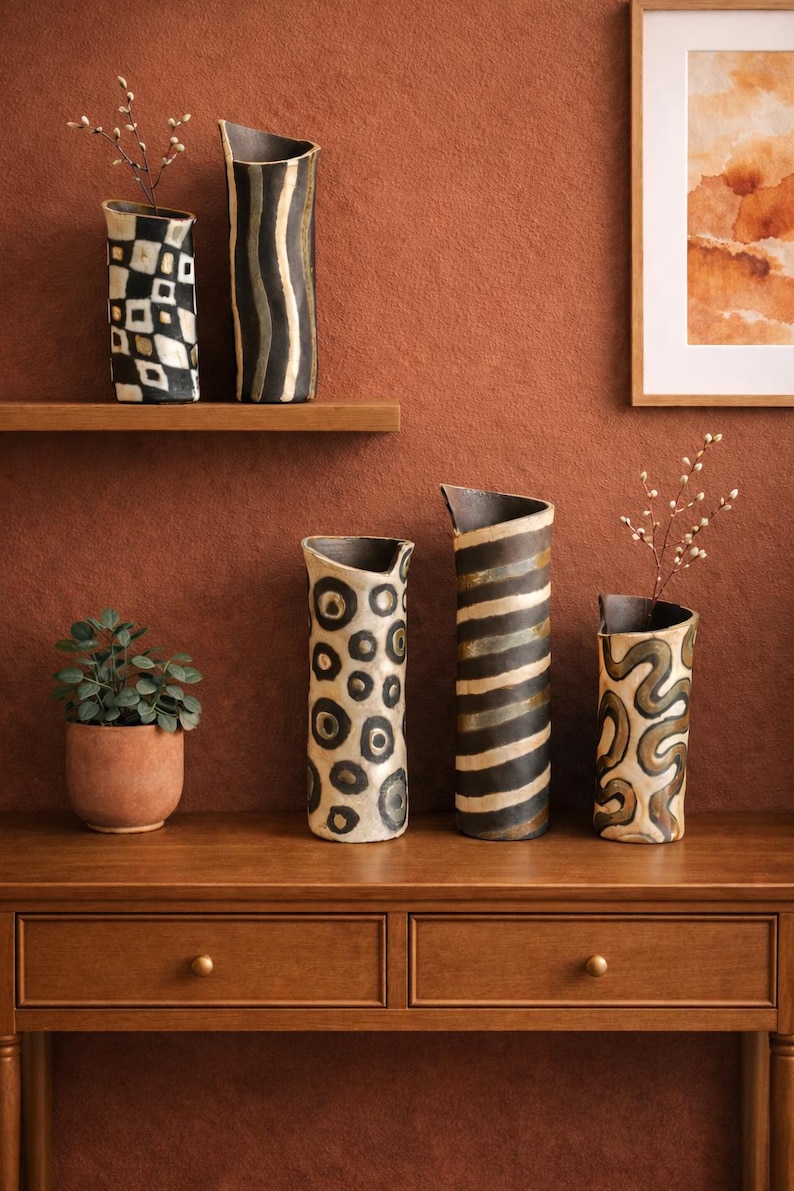 May include: A collection of decorative ceramic vases with unique patterns. The vases feature designs in black, white, and brown, including geometric shapes, stripes, and organic swirls. Some vases hold small branches with delicate white flowers.