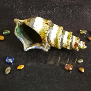 Puede incluir: Una concha de cer&aacute;mica con rayas verdes, blancas y marrones, sobre una superficie oscura. Alrededor de la concha hay peque&ntilde;as piedras de vidrio de colores en tonos azules, dorados y verdes. El interior de la concha es verde oscuro.