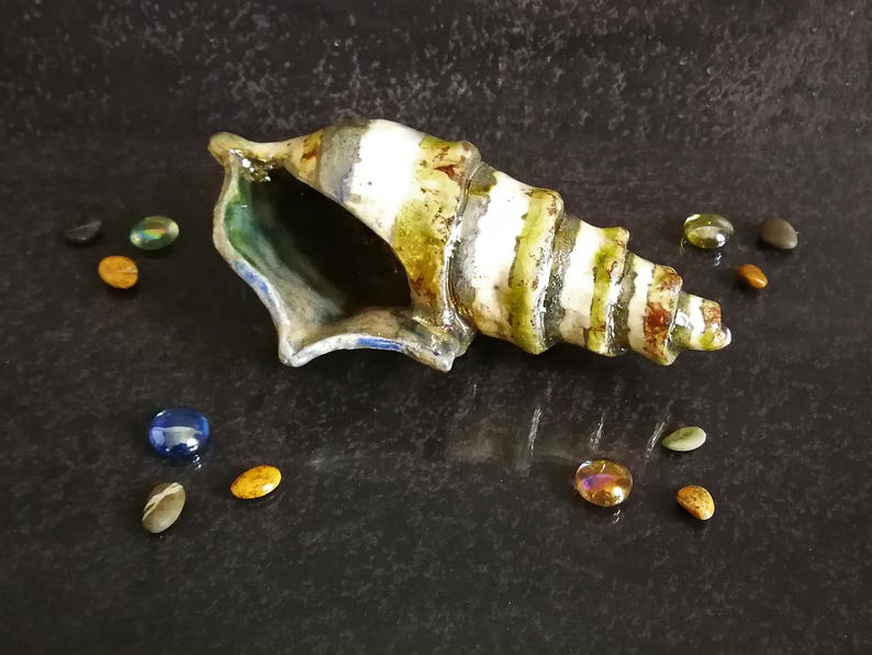 Puede incluir: Una concha de cer&aacute;mica con rayas verdes, blancas y marrones, sobre una superficie oscura. Alrededor de la concha hay peque&ntilde;as piedras de vidrio de colores en tonos azules, dorados y verdes. El interior de la concha es verde oscuro.