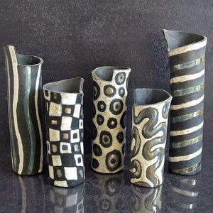 May include: Five ceramic vases with unique designs. The vases are cylindrical with asymmetrical tops. Patterns include vertical stripes, wavy lines, checkerboard, and circles. The color palette is primarily black, white, and gray.