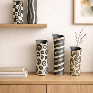 May include: A collection of ceramic vases with unique patterns. The vases feature designs such as geometric shapes, stripes, and organic swirls in black and white. Some vases have a curved top and are displayed on a wooden shelf and cabinet.