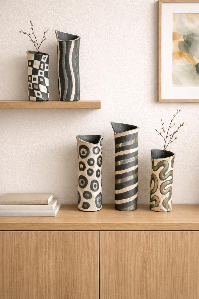 May include: A collection of ceramic vases with unique patterns. The vases feature designs such as geometric shapes, stripes, and organic swirls in black and white. Some vases have a curved top and are displayed on a wooden shelf and cabinet.