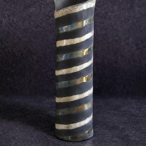 May include: A tall, cylindrical ceramic vase with a dark gray exterior. The vase features alternating black, white, and gold horizontal stripes. The top edge is curved and angled. The vase is approximately 12 inches tall.
