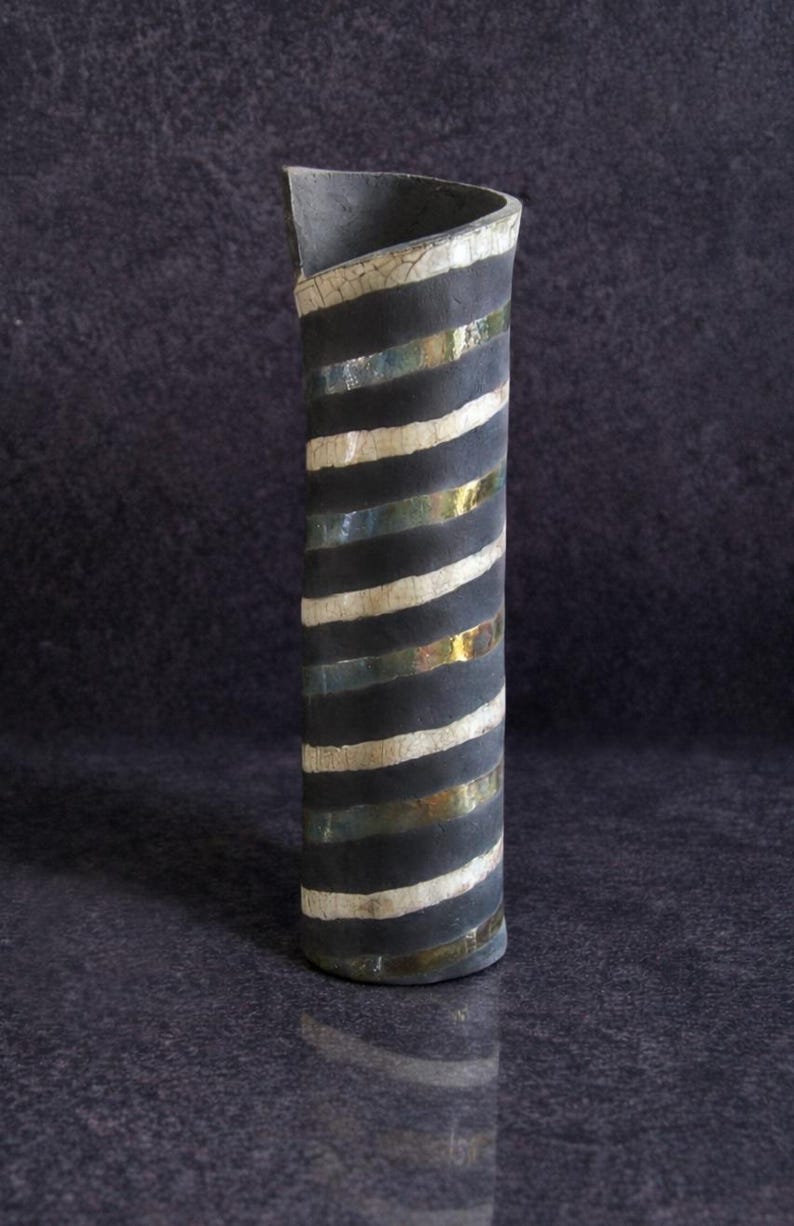 May include: A tall, cylindrical ceramic vase with a dark gray exterior. The vase features alternating black, white, and gold horizontal stripes. The top edge is curved and angled. The vase is approximately 12 inches tall.