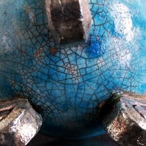 May include: A close-up of a ceramic vase with a turquoise crackle glaze. The vase has a rounded shape and is supported by three silver-colored feet. The glaze has a network of fine cracks.