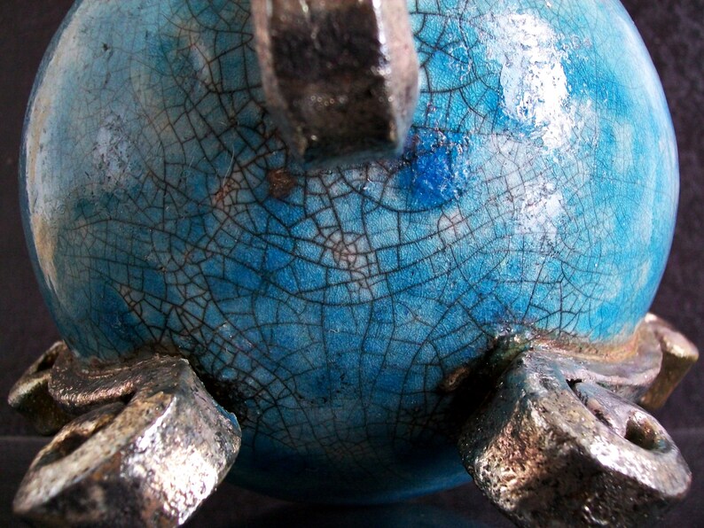 May include: A close-up of a ceramic vase with a turquoise crackle glaze. The vase has a rounded shape and is supported by three silver-colored feet. The glaze has a network of fine cracks.