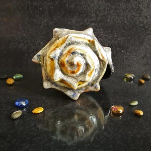 Puede incluir: Un anillo de cer&aacute;mica con forma de concha marina con un dise&ntilde;o en espiral en tonos blancos, marrones y azules. El anillo est&aacute; rodeado de peque&ntilde;as piedras de colores. Una joya &uacute;nica.