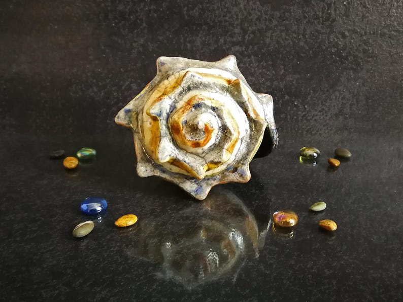 Puede incluir: Un anillo de cer&aacute;mica con forma de concha marina con un dise&ntilde;o en espiral en tonos blancos, marrones y azules. El anillo est&aacute; rodeado de peque&ntilde;as piedras de colores. Una joya &uacute;nica.