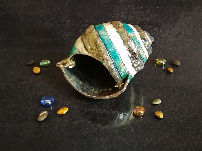 Puede incluir: Una concha decorativa con rayas alternas blancas, turquesas y grises oscuros. Peque&ntilde;os guijarros de vidrio de colores rodean la concha sobre una superficie oscura y reflectante. El interior de la concha es oscuro.