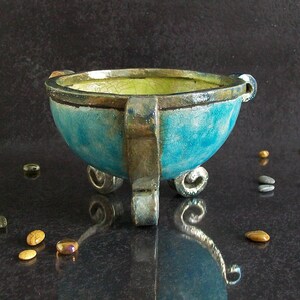 May include: A handcrafted, turquoise-colored ceramic bowl with a textured surface and a light green interior. The bowl is supported by three ornate, silver-colored legs and features decorative handles. Small, colorful stones are scattered around the base.