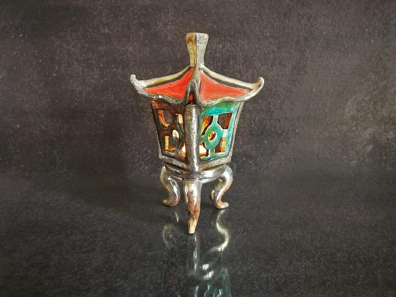 May include: A decorative silver-colored lantern with a red roof and green and brown accents. The lantern has a square shape with ornate cutouts and sits on three curved legs. The lantern's design is reminiscent of Asian architecture.
