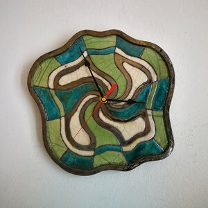 Pu&ograve; includere: Un orologio da parete con una cornice in ceramica dai bordi irregolari e ondulati, caratterizzata da una finitura smaltata a craquel&eacute;. Il quadrante presenta un motivo vorticoso e segmentato in tonalit&agrave; verde acqua, verde chiaro e crema, con lancette nere e rosse.