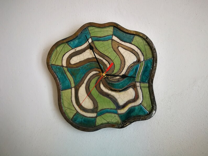 Pu&ograve; includere: Un orologio da parete con una cornice in ceramica dai bordi irregolari e ondulati, caratterizzata da una finitura smaltata a craquel&eacute;. Il quadrante presenta un motivo vorticoso e segmentato in tonalit&agrave; verde acqua, verde chiaro e crema, con lancette nere e rosse.