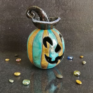 May include: A ceramic jack-o'-lantern featuring vertical stripes in alternating shades of teal and bronze. The pumpkin has a carved face with cutouts for eyes and a mouth, topped with a glossy black, curled stem that resembles a witch's hat.