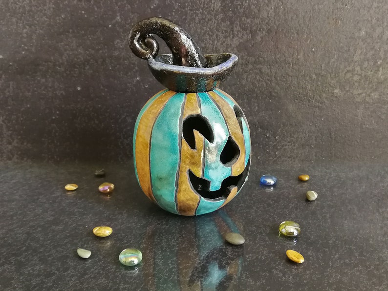 May include: A ceramic jack-o'-lantern featuring vertical stripes in alternating shades of teal and bronze. The pumpkin has a carved face with cutouts for eyes and a mouth, topped with a glossy black, curled stem that resembles a witch's hat.