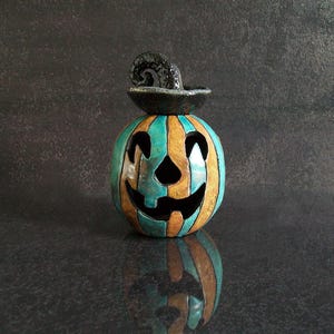 May include: A ceramic jack-o'-lantern featuring alternating vertical stripes of teal and tan glaze. The pumpkin has a carved face with triangular eyes and a wide, jagged mouth. A black, textured, curled stem sits atop the pumpkin.