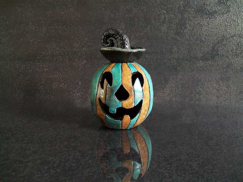 May include: A ceramic jack-o'-lantern featuring alternating vertical stripes of teal and tan glaze. The pumpkin has a carved face with triangular eyes and a wide, jagged mouth. A black, textured, curled stem sits atop the pumpkin.