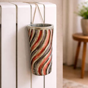 May include: A cylindrical vase with a spiral design of red, gray, and black stripes. The vase is suspended by a natural-colored cord and hangs against a white surface. The vase has a metallic rim.
