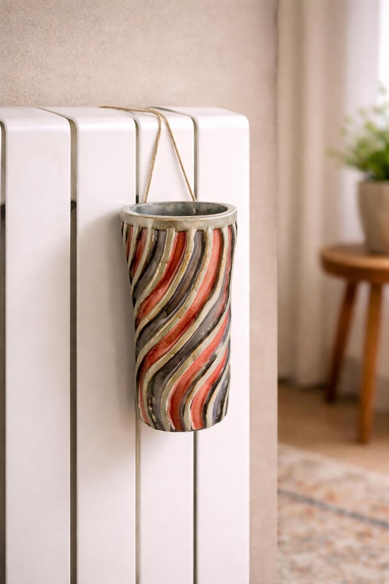 May include: A cylindrical vase with a spiral design of red, gray, and black stripes. The vase is suspended by a natural-colored cord and hangs against a white surface. The vase has a metallic rim.
