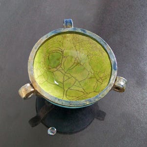 May include: A handmade ceramic bowl with a vibrant green interior featuring a crackle glaze design. The bowl has a blue rim and three small handles. The bowl is sitting on a dark surface.