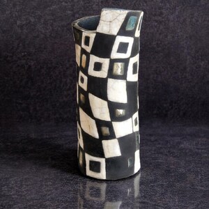 May include: A tall, cylindrical vase with a black and white checkered pattern. The ceramic vase has a unique shape with a curved top and a glossy finish. The design incorporates squares and rectangles, creating a striking visual effect.