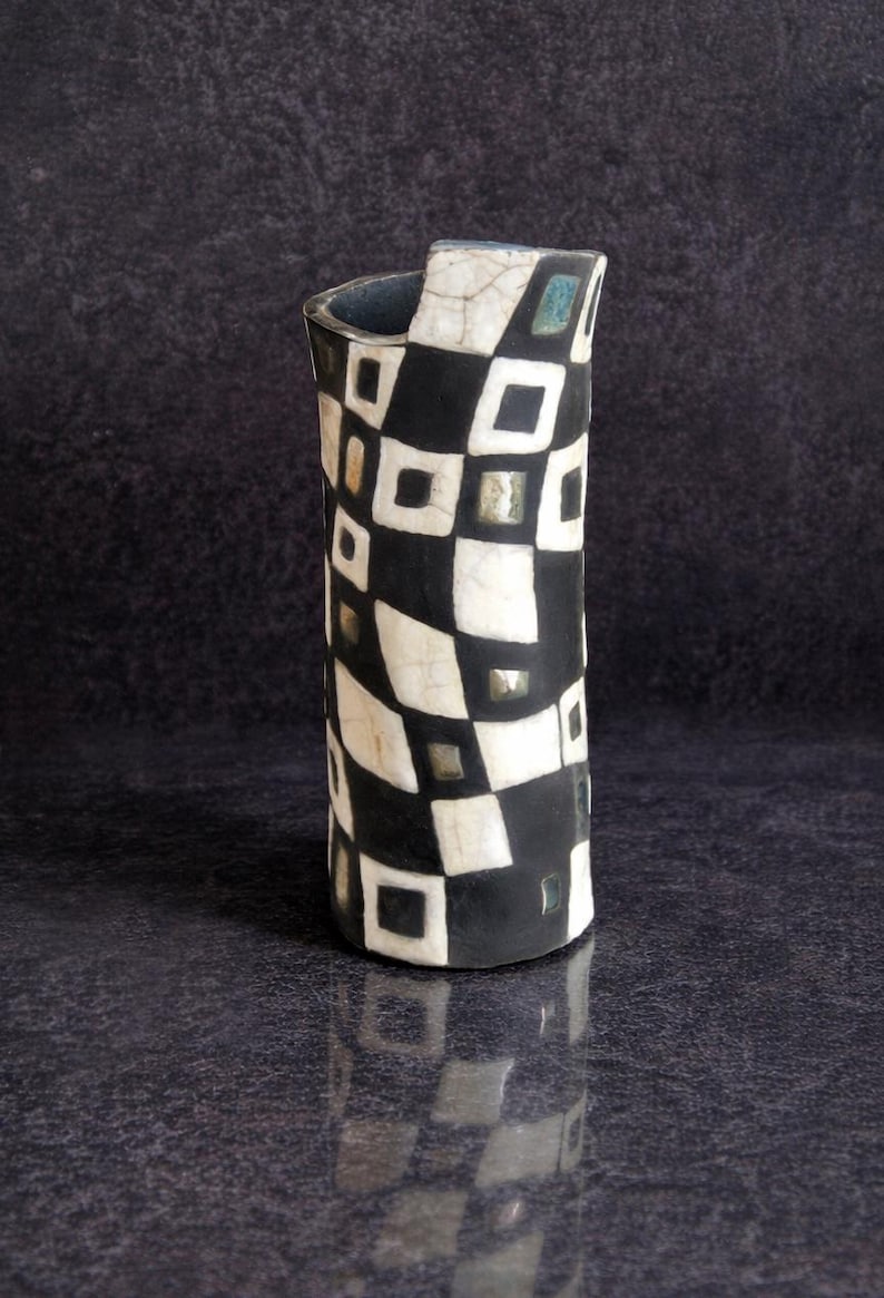 May include: A tall, cylindrical vase with a black and white checkered pattern. The ceramic vase has a unique shape with a curved top and a glossy finish. The design incorporates squares and rectangles, creating a striking visual effect.