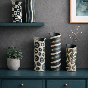 May include: A collection of ceramic vases with unique patterns, including geometric shapes, stripes, and organic designs. The vases are in shades of white, black, and gray, displayed on a teal console table and shelf. A small potted plant and framed artwork complete the decor.